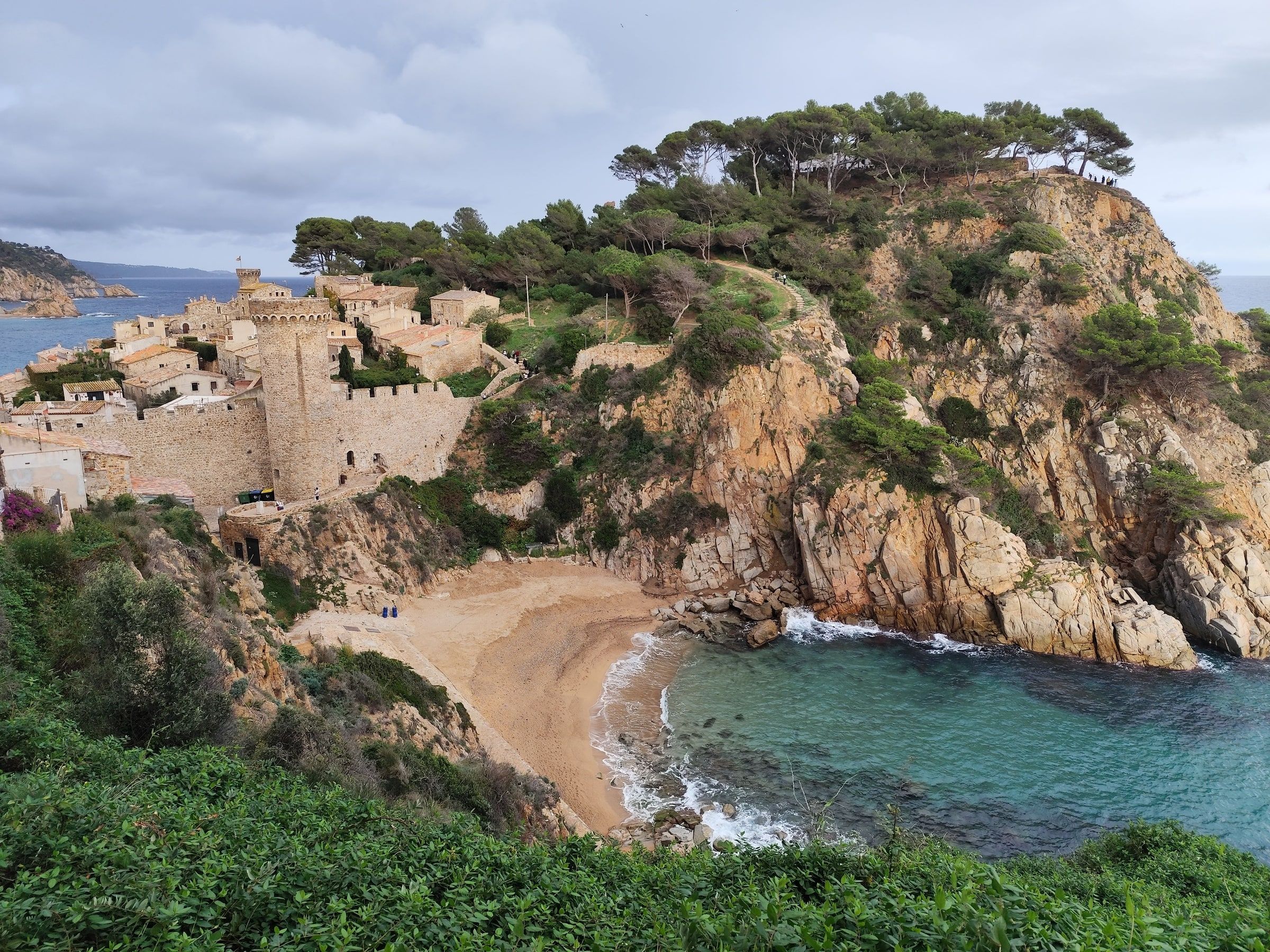 Tossa de Mar uppifrån: gamla stan med muren och tornen, en liten sandstrand och turkost vatten mellan klippor med tallar.