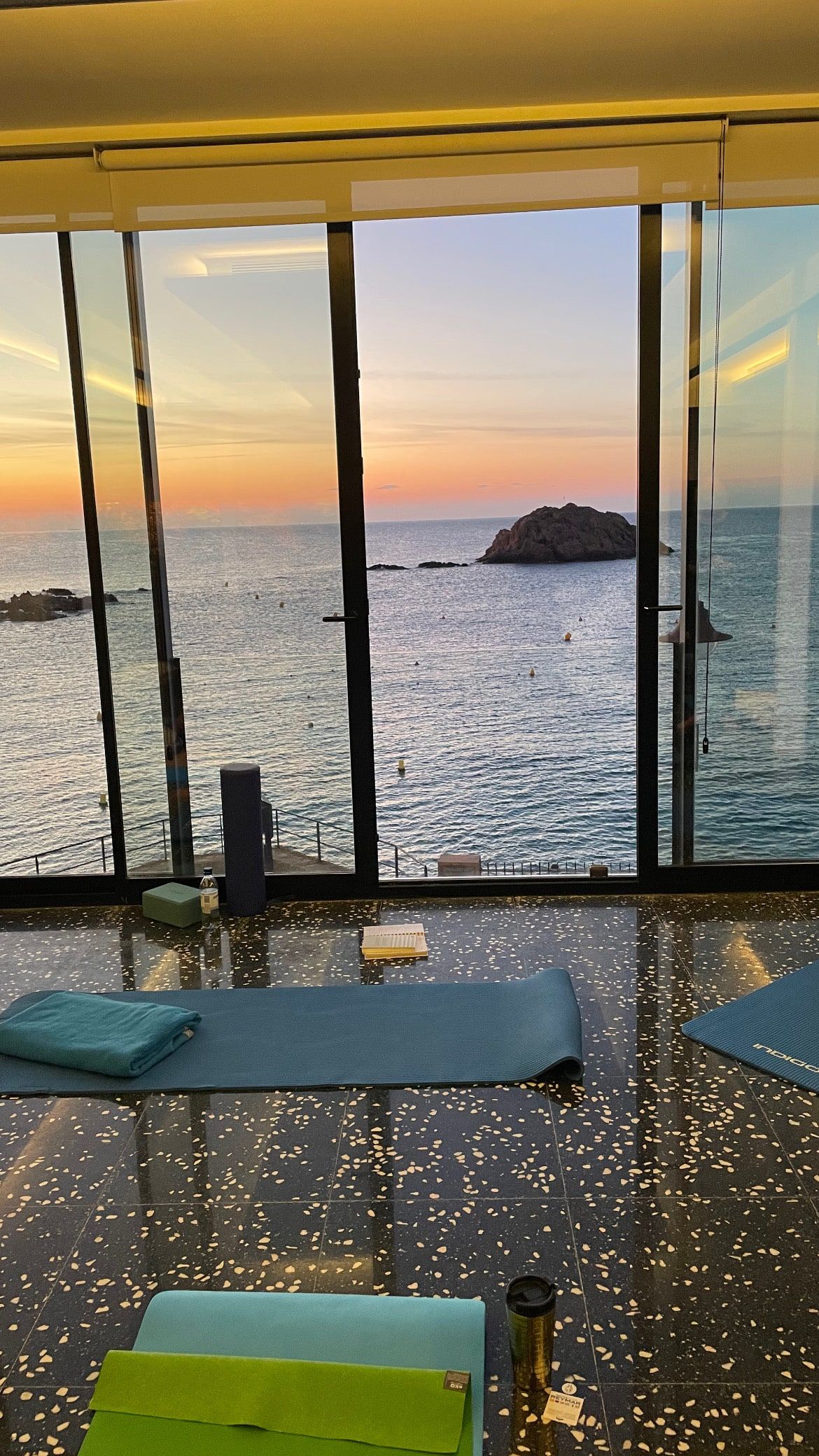 Yogasal inomhus med mattor framför glasväggar mot havet och klippan i soluppgången.