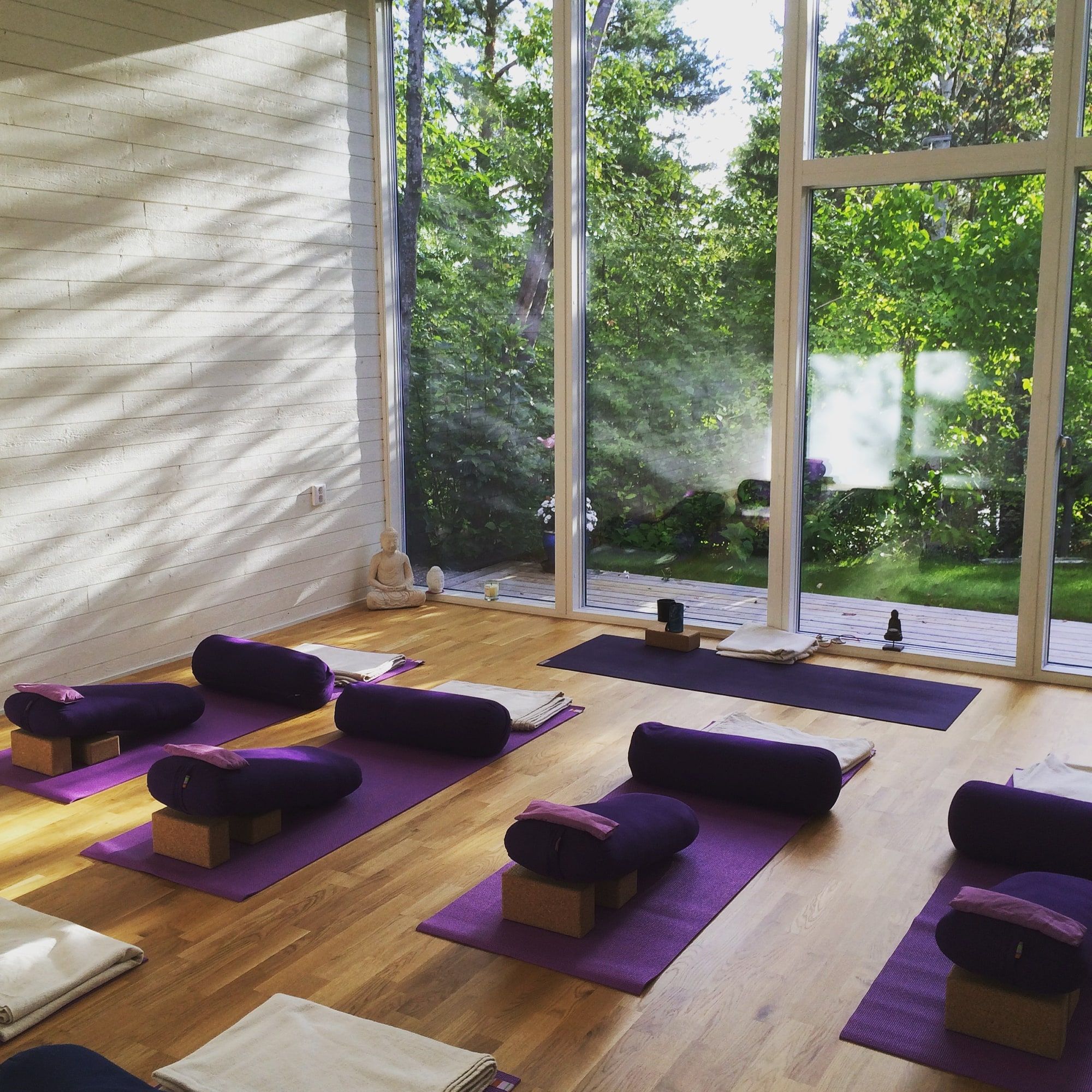 Yogasalen i morgonljus med ekgolv, lila mattor och bolster och glasväggar mot grönskan.